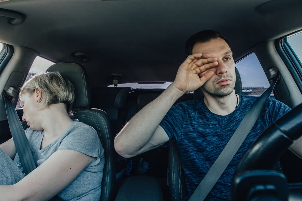 Man and Woman in a car, struggling to stay awake and yawn while holding the wheel, illustrating driver fatigue and drowsy driving.