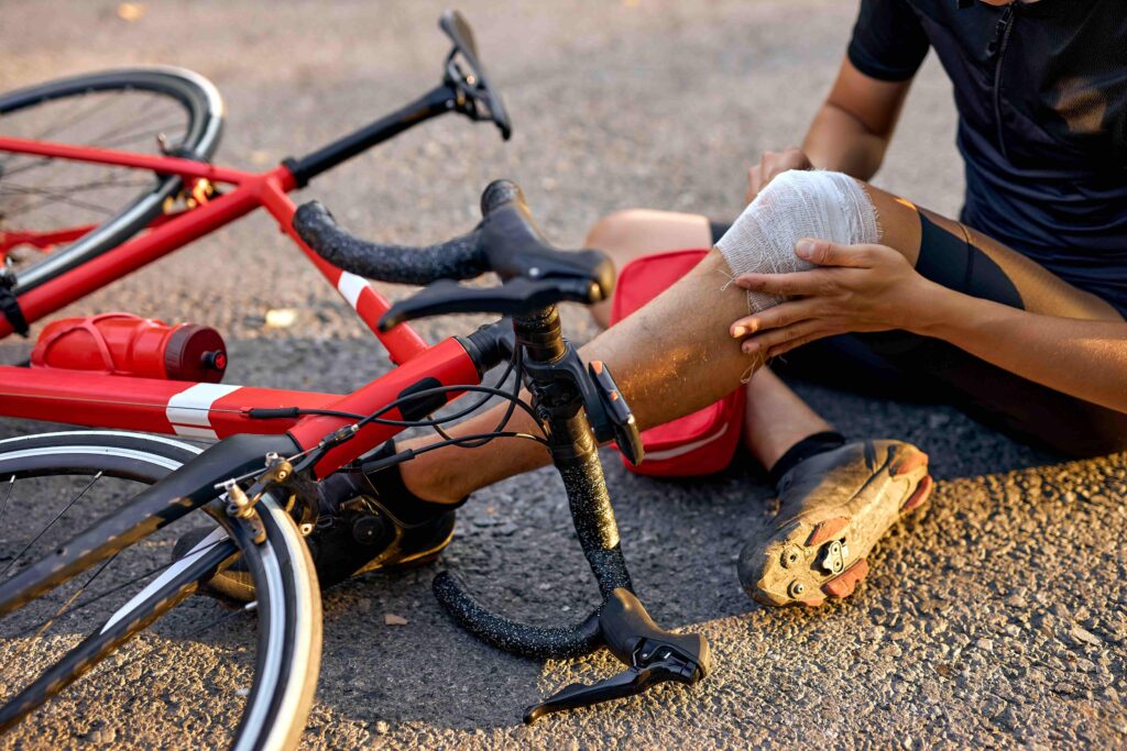 Injured cyclist sitting on road beside fallen red bicycle, holding bandaged knee after bike accident.
