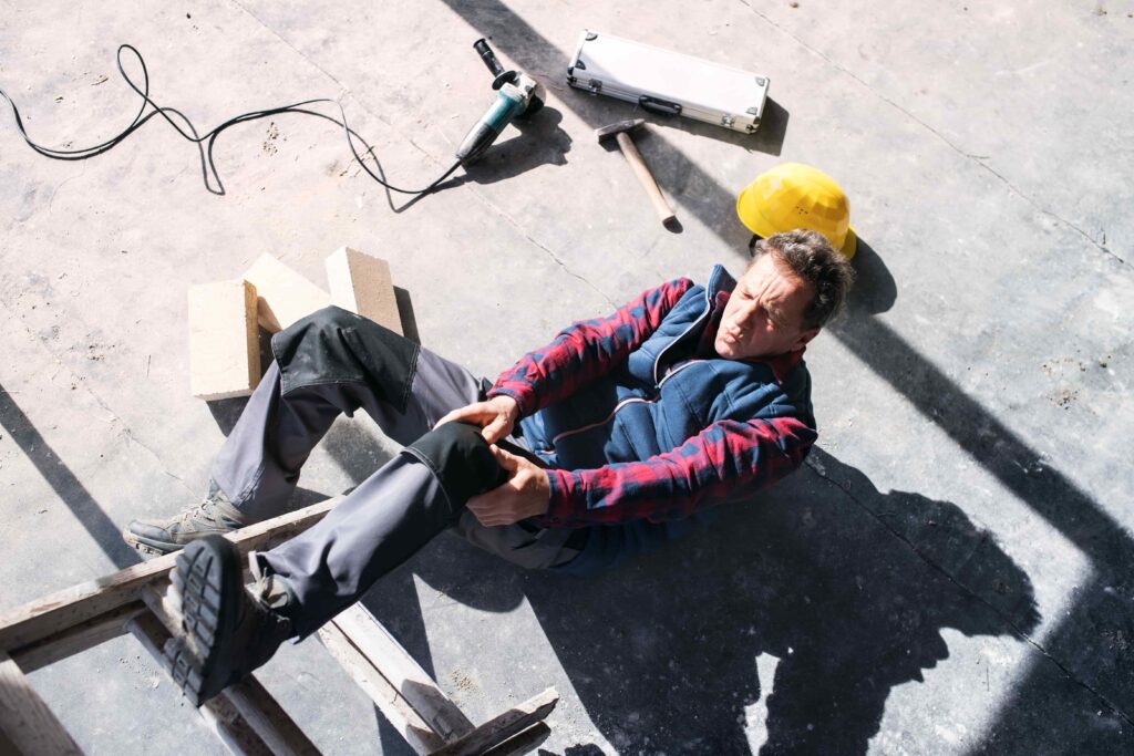 Injured construction worker sitting on floor holding knee after workplace fall, with tools scattered nearby.